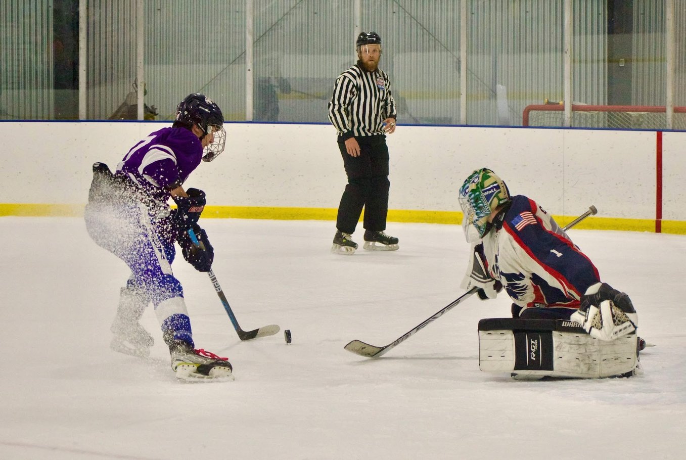 High school ice hockey player in purple jersey driving to the net during a competitive game, battling the goalie in a one-on-one breakaway while a referee observes — showcasing elite stickhandling skills, shooting accuracy, speed, edge control, and game-ready performance for college hockey recruiting scouts and NCAA coaches.